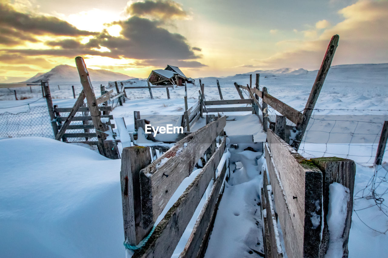 WOODEN POSTS ON SNOW COVERED FIELD AGAINST SKY