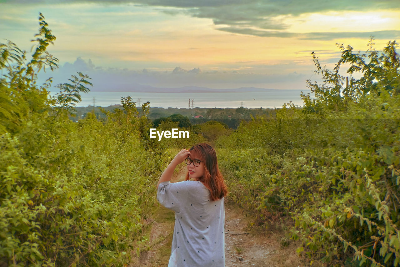 Rear view portrait of woman standing amidst plants against sky during sunset