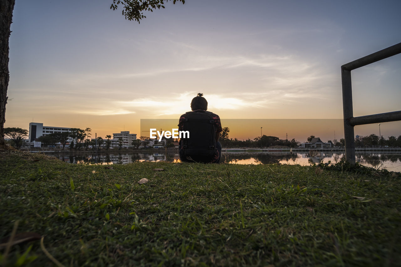 Rear view of man sitting on field against sky during sunset