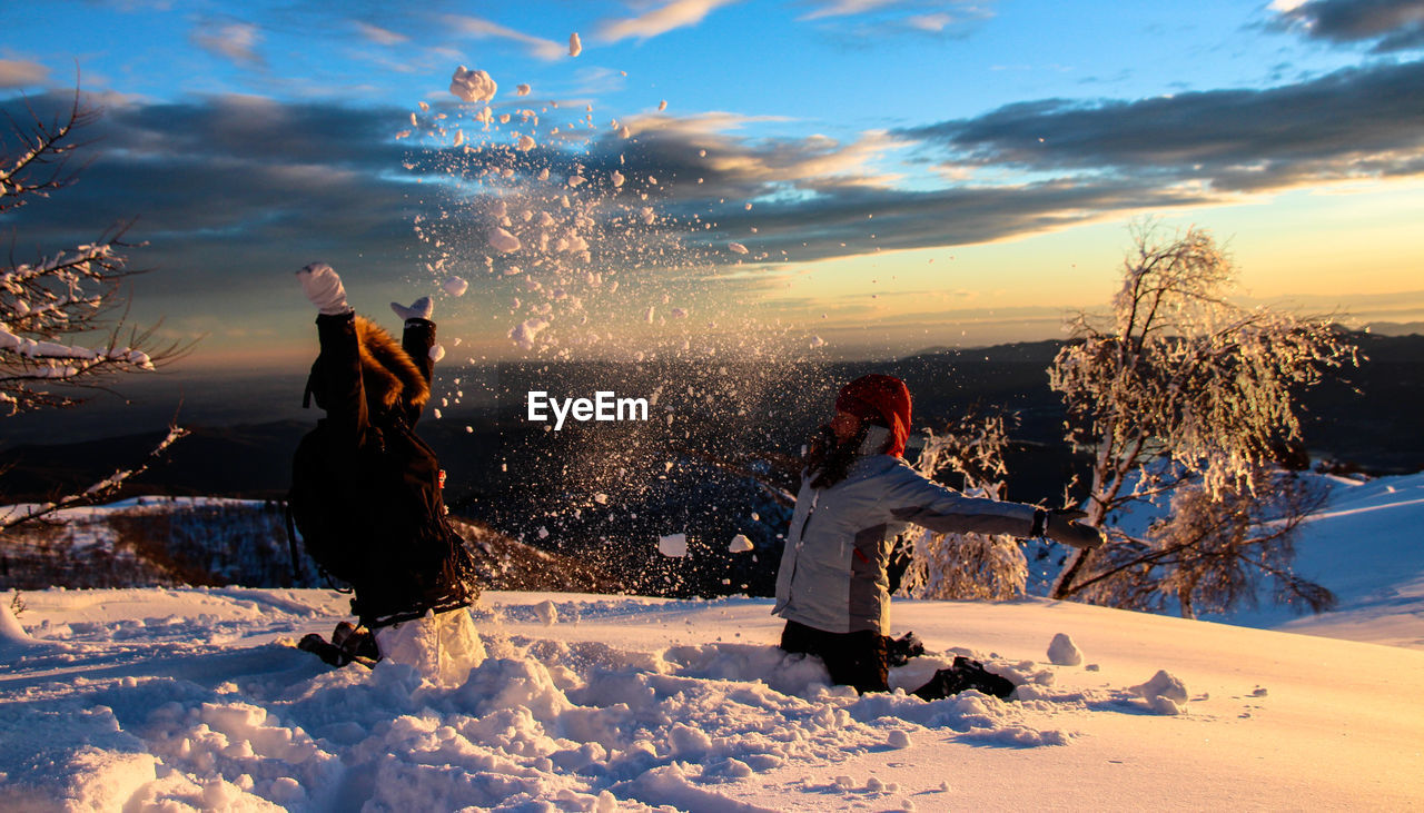 Kids throwing snow against cloudy sky during sunset