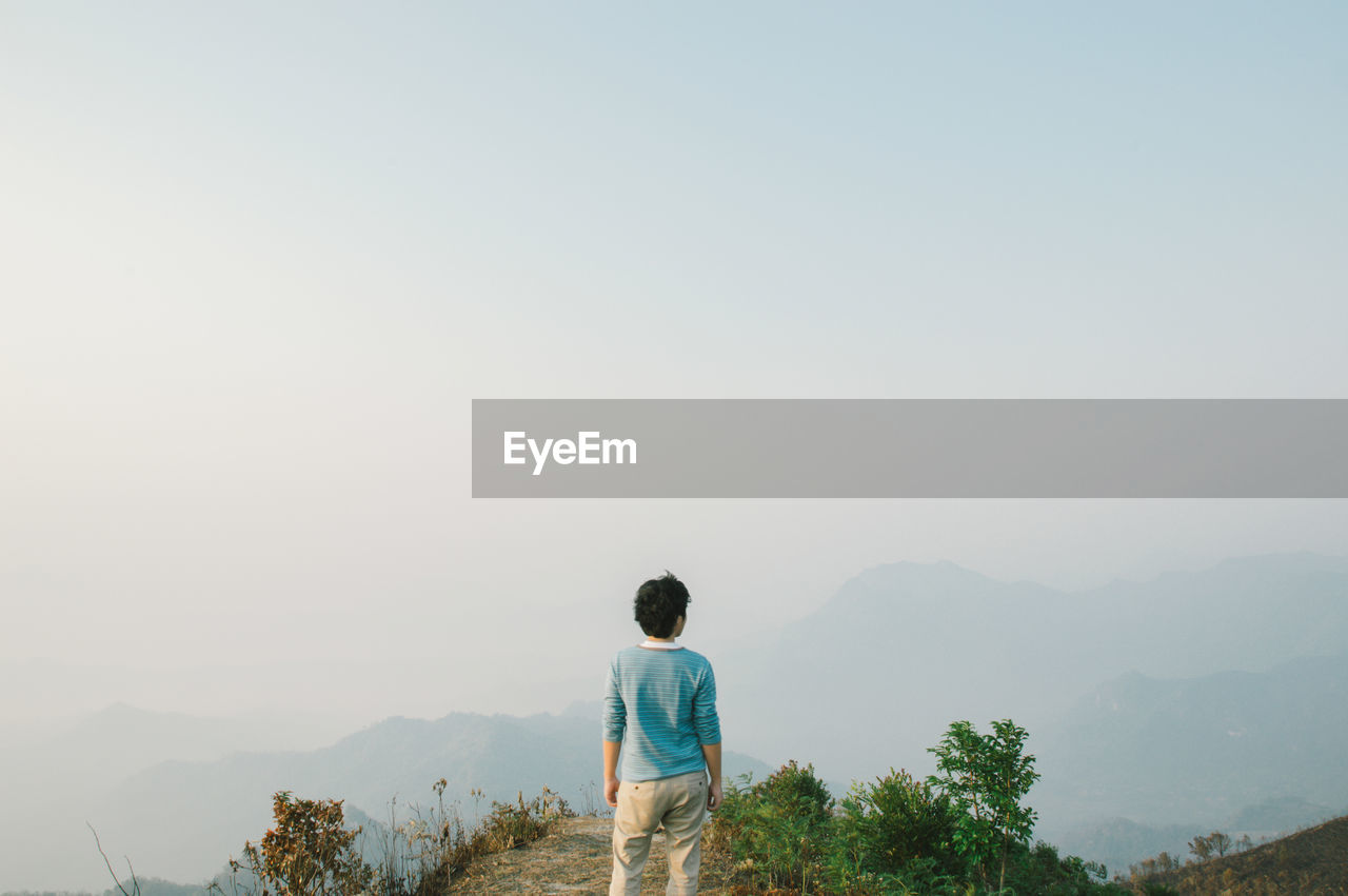 Rear view of man standing on cliff against sky in foggy weather