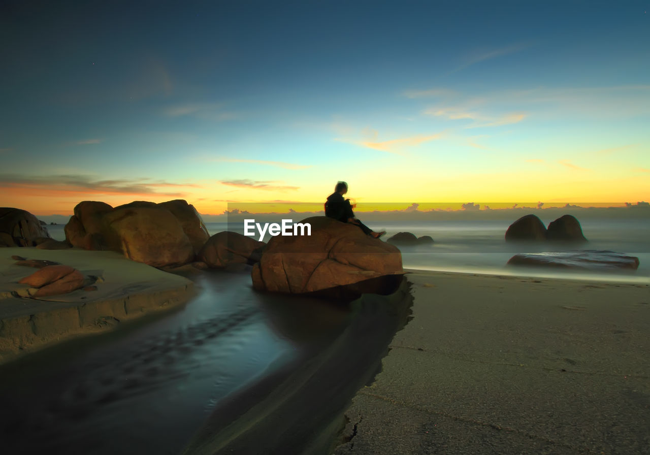 Woman sitting on rock at beach against sky during sunrise