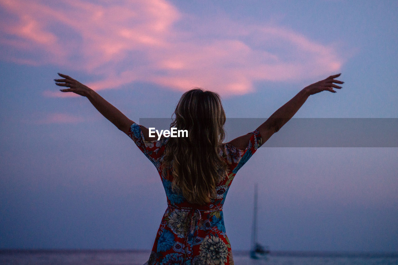Rear view of woman with arms raised against sky during sunset