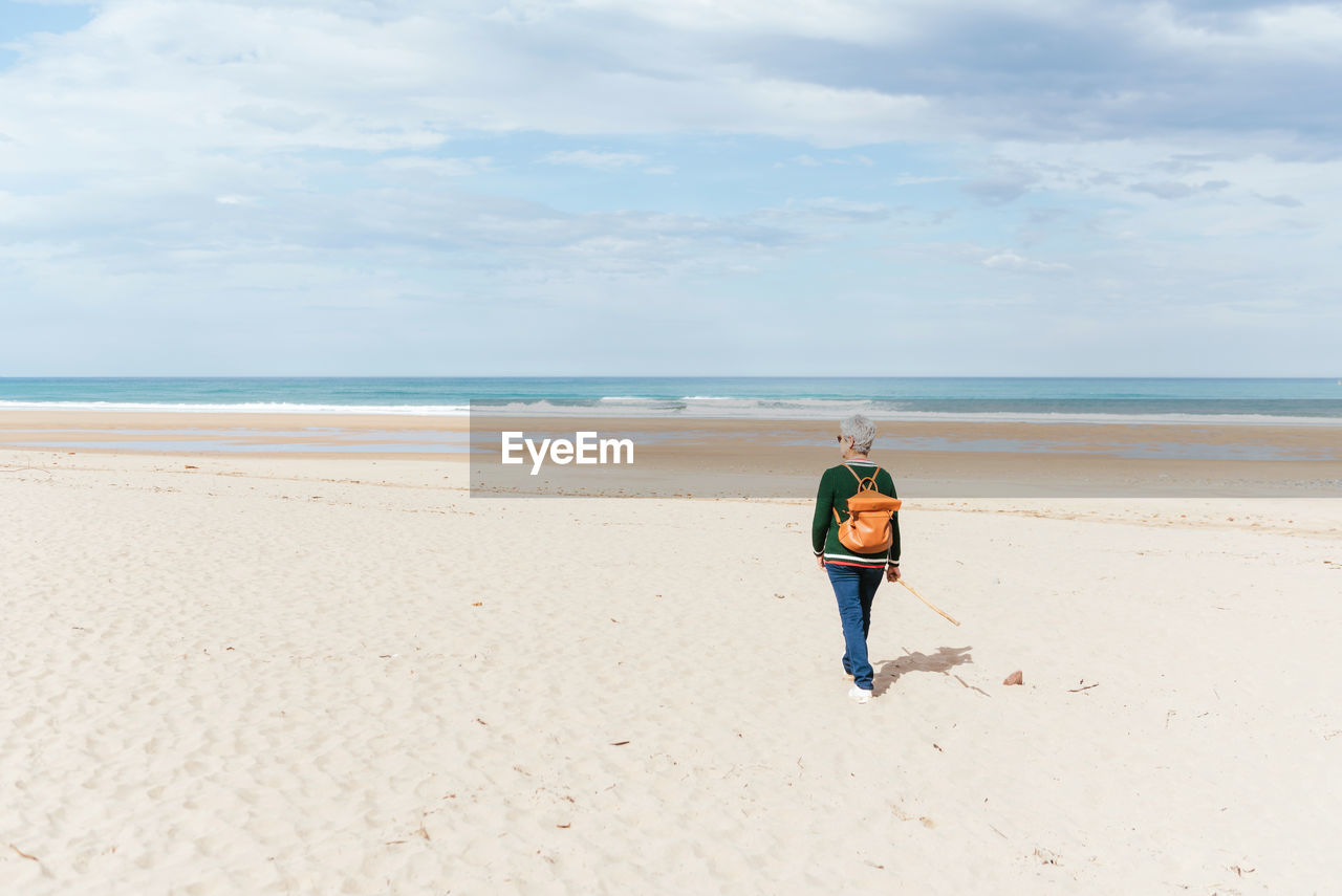 Back view of unrecognizable senior female trekker walking on sandy shore against endless sea during trip