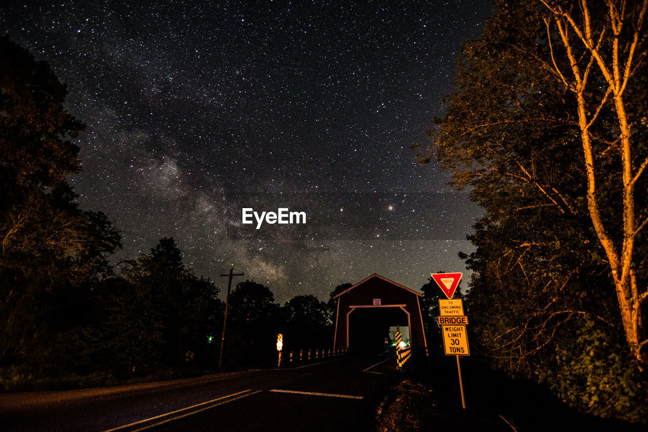 Sign board by road leading towards bridge against star field at night