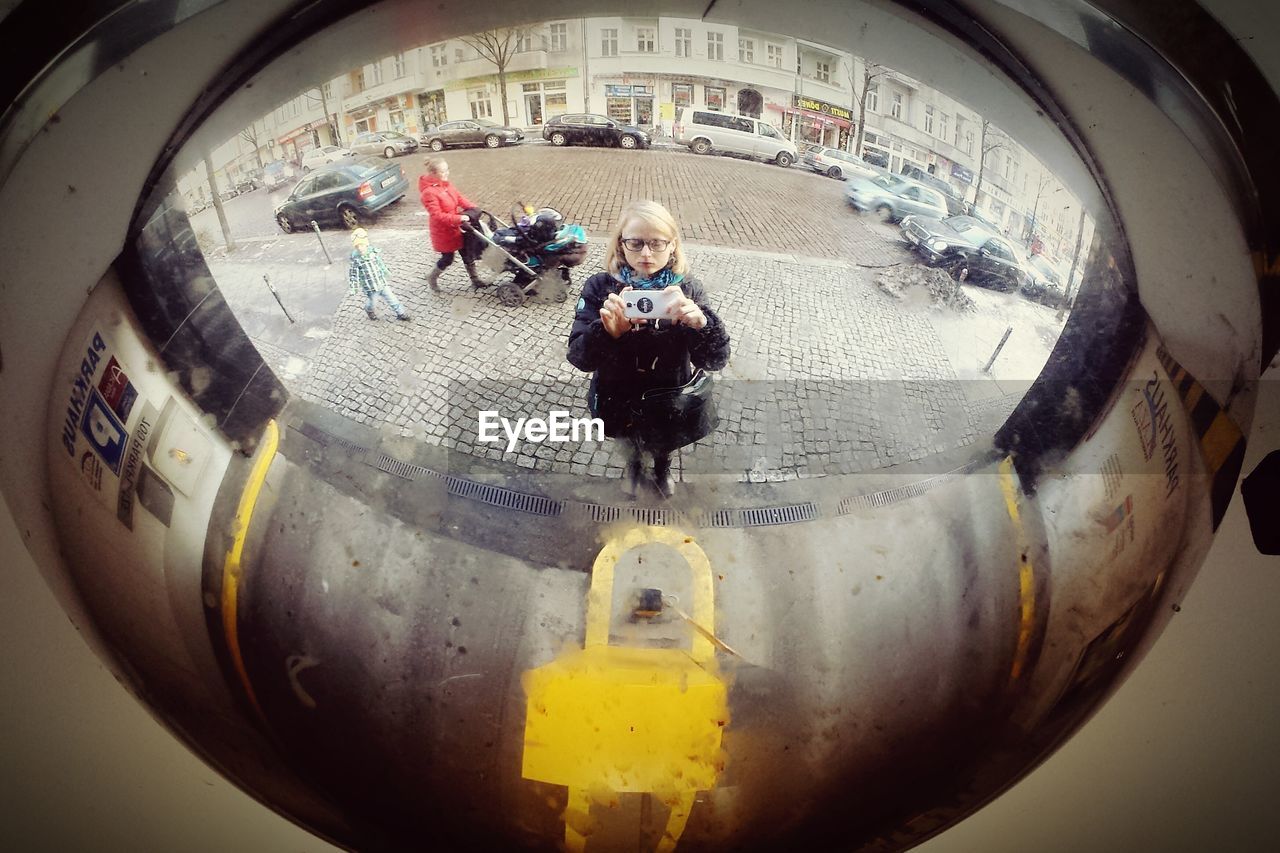 Woman taking selfie on mirror against buildings and cars in city