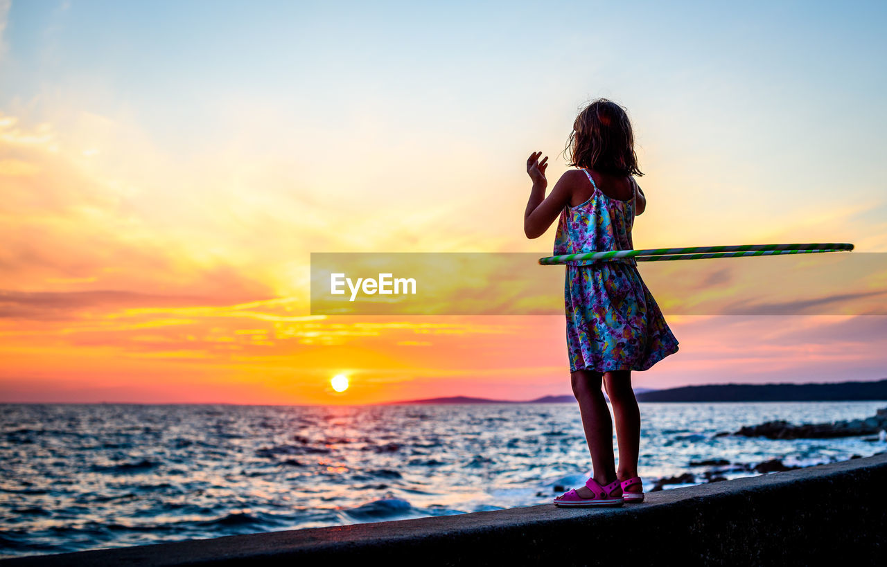 Full length of girl playing with hula hoop on shore at beach against sky during sunset