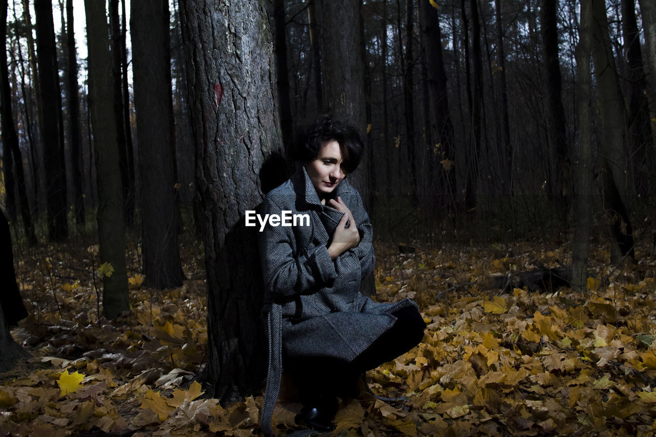 Woman sitting by tree trunk in forest during autumn