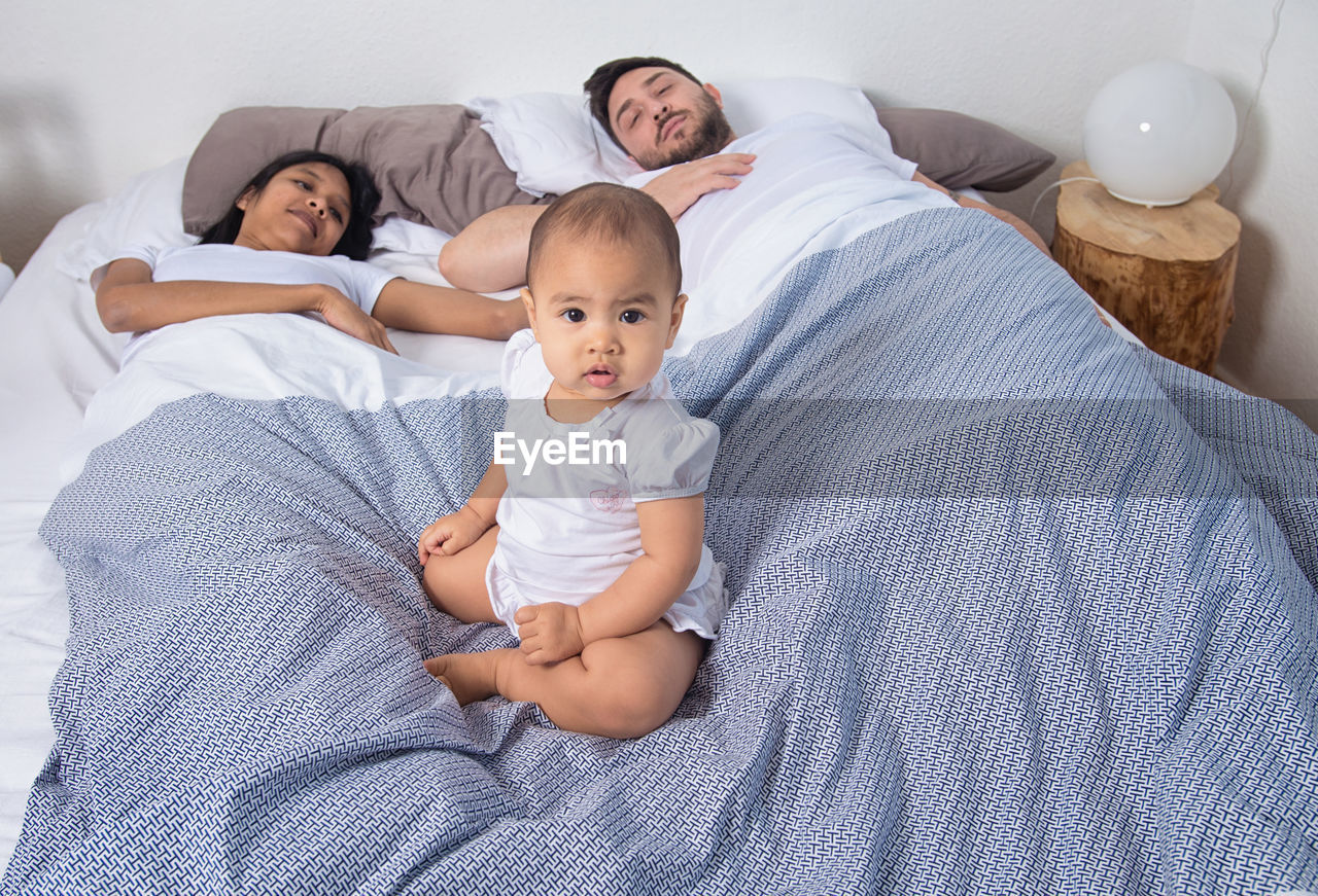 High angle view of baby sitting amidst parents sleeping on bed at home