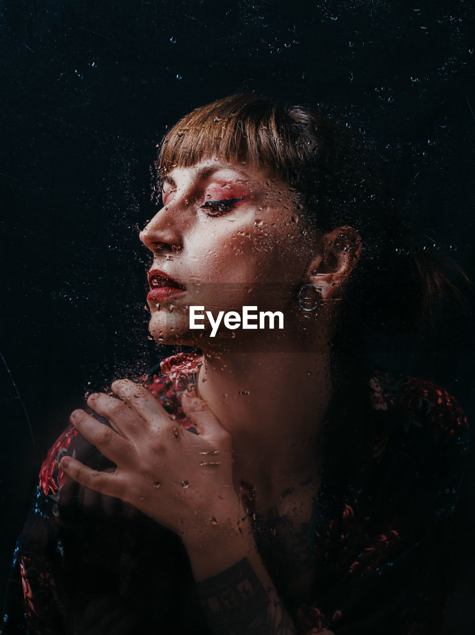 Unemotional female with arm tattoo standing behind translucent glass with water droplets