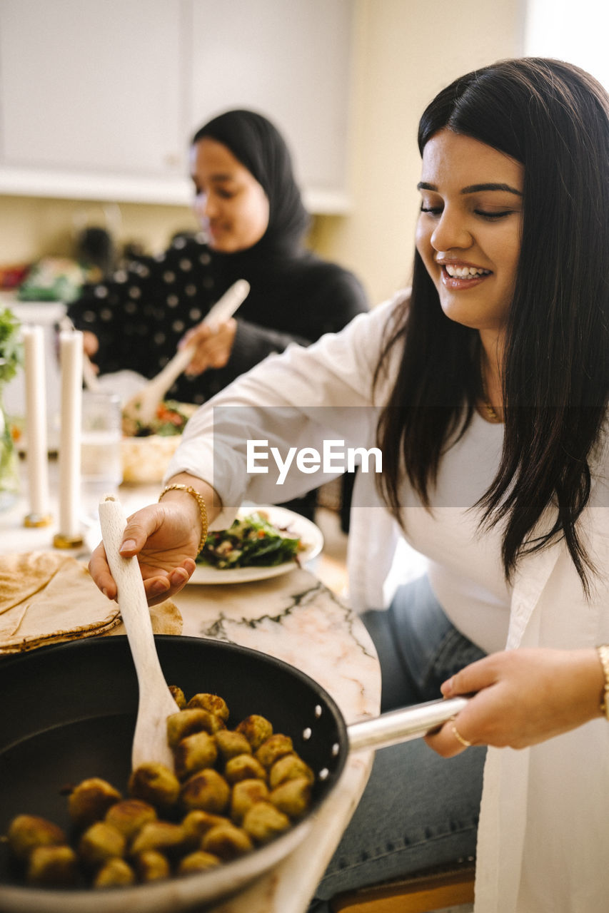 Smiling young woman serving food from cooking pan while sitting at dining table