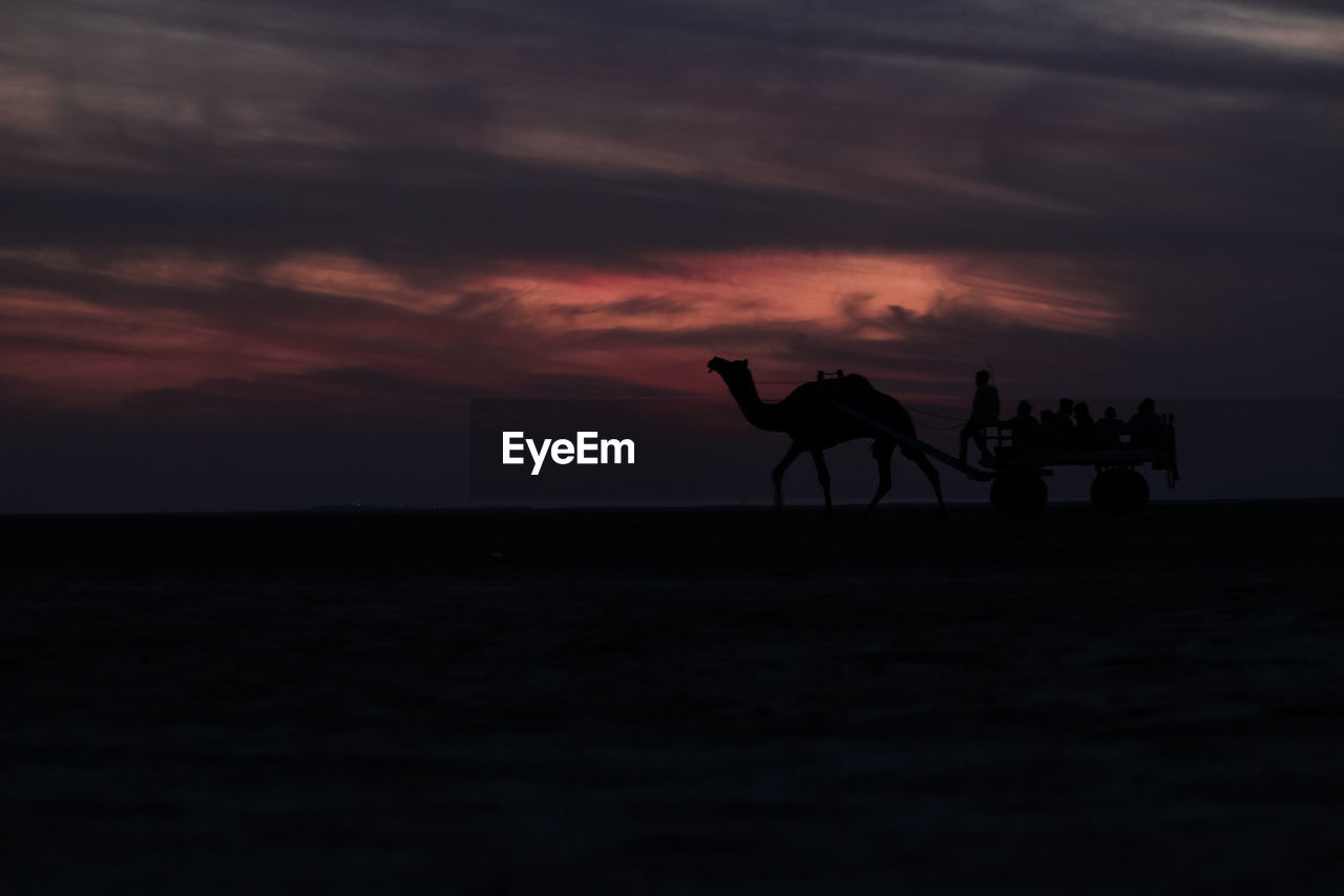 SILHOUETTE PEOPLE RIDING MOTORCYCLE ON BEACH AGAINST SKY DURING SUNSET