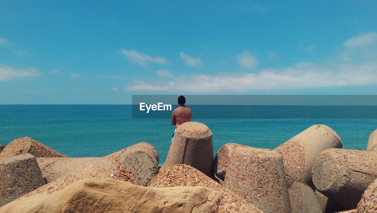 Rear view of shirtless man on tetrapods at sea shore against sky