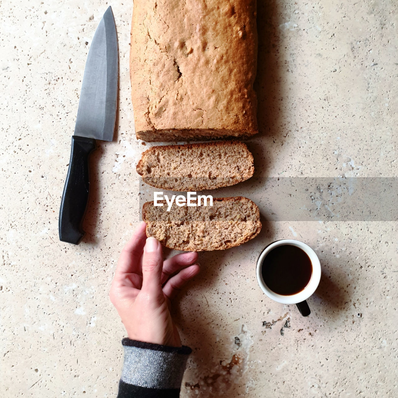 High angle view of hand holding slice of bread with coffee and knife on stone table