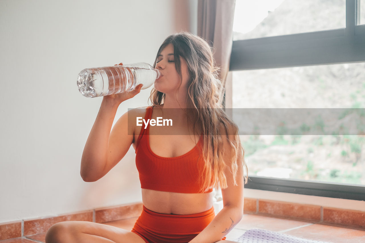 portrait of young woman drinking water while standing against wall
