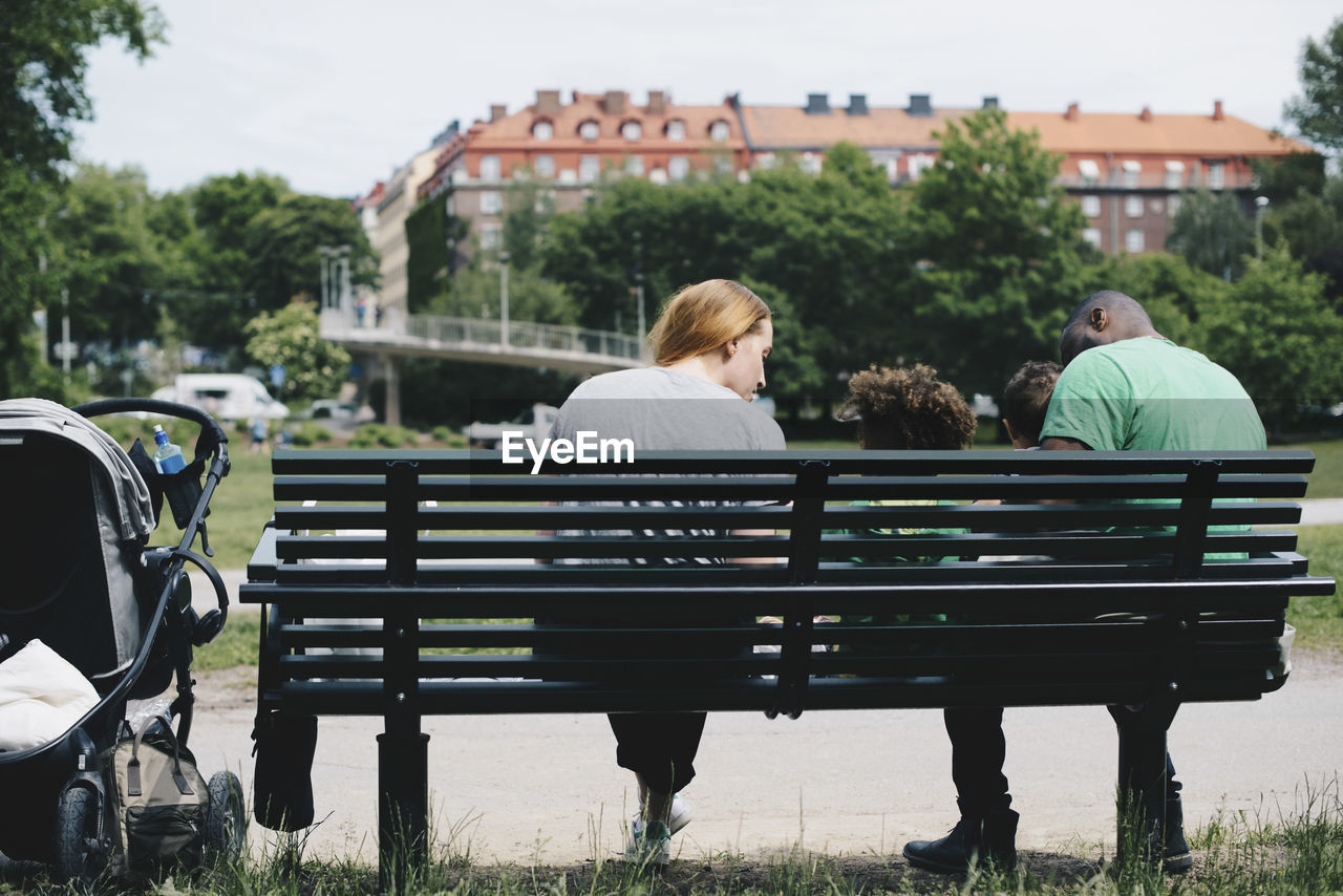 Rear view of family sitting on bench at park in city