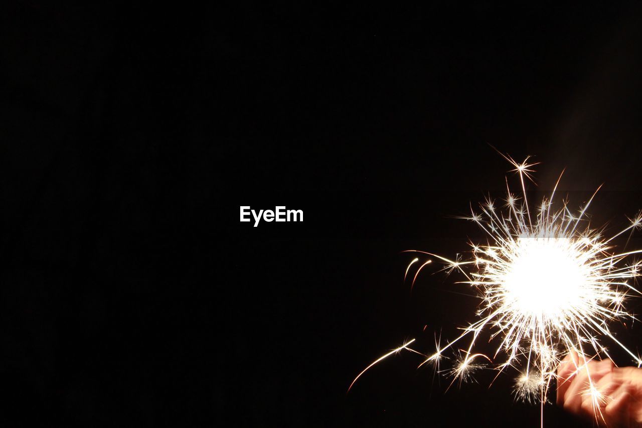 Cropped hand holding lit sparkler against black background