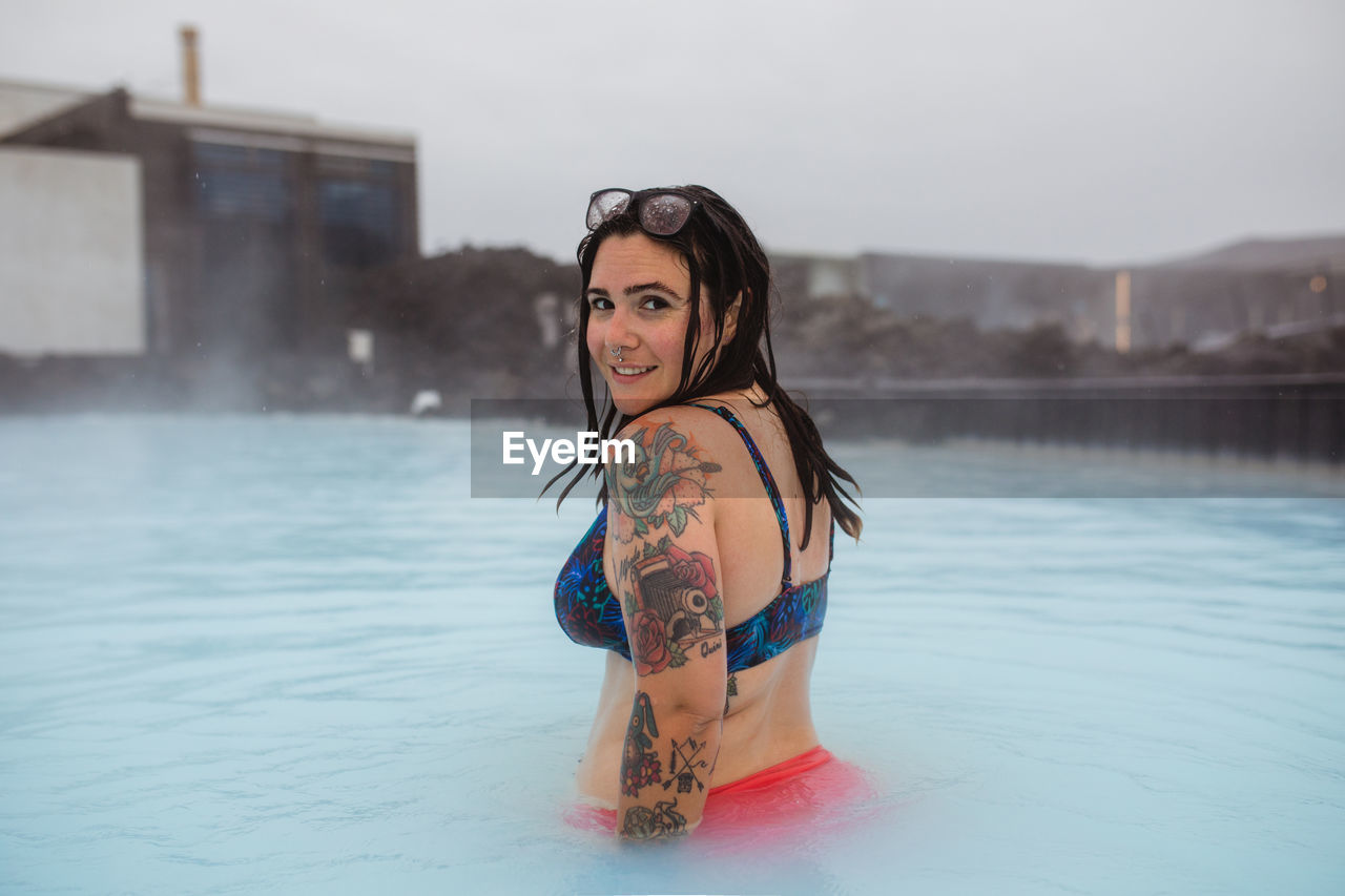 Side view of young tattooed hipster in swimsuit posing in blue water between rocks