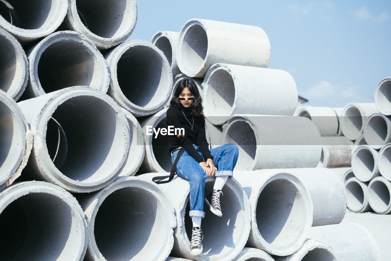 FULL LENGTH PORTRAIT OF BOY STANDING ON STACK OF PIPES AGAINST SKY