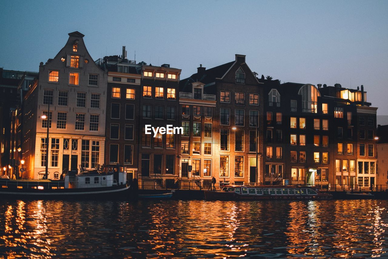 ILLUMINATED BUILDINGS BY RIVER AGAINST SKY AT DUSK