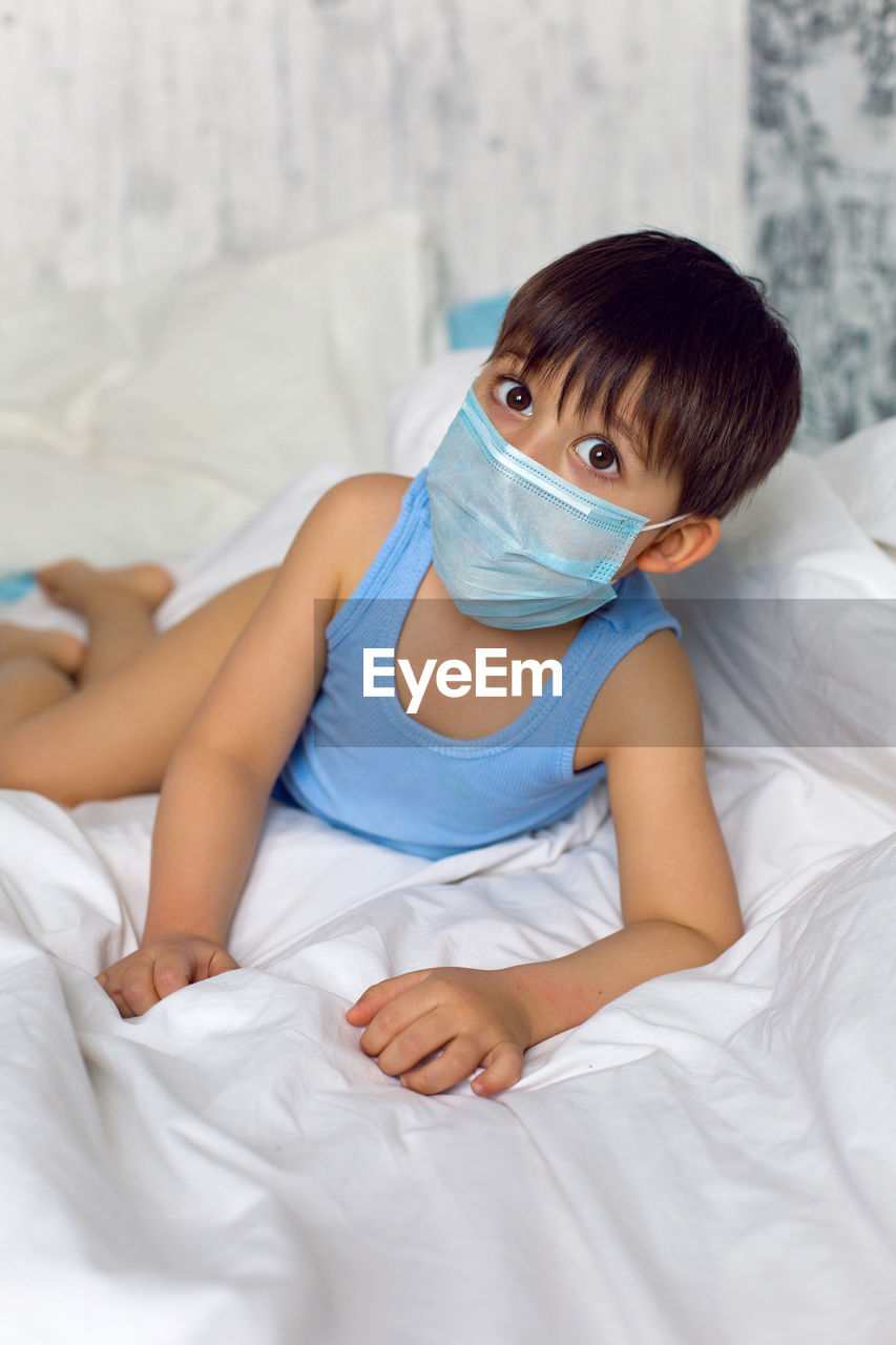 Boy in a blue t-shirt and a medical mask sits on a bed in quarantine during the covid-19 epidemic