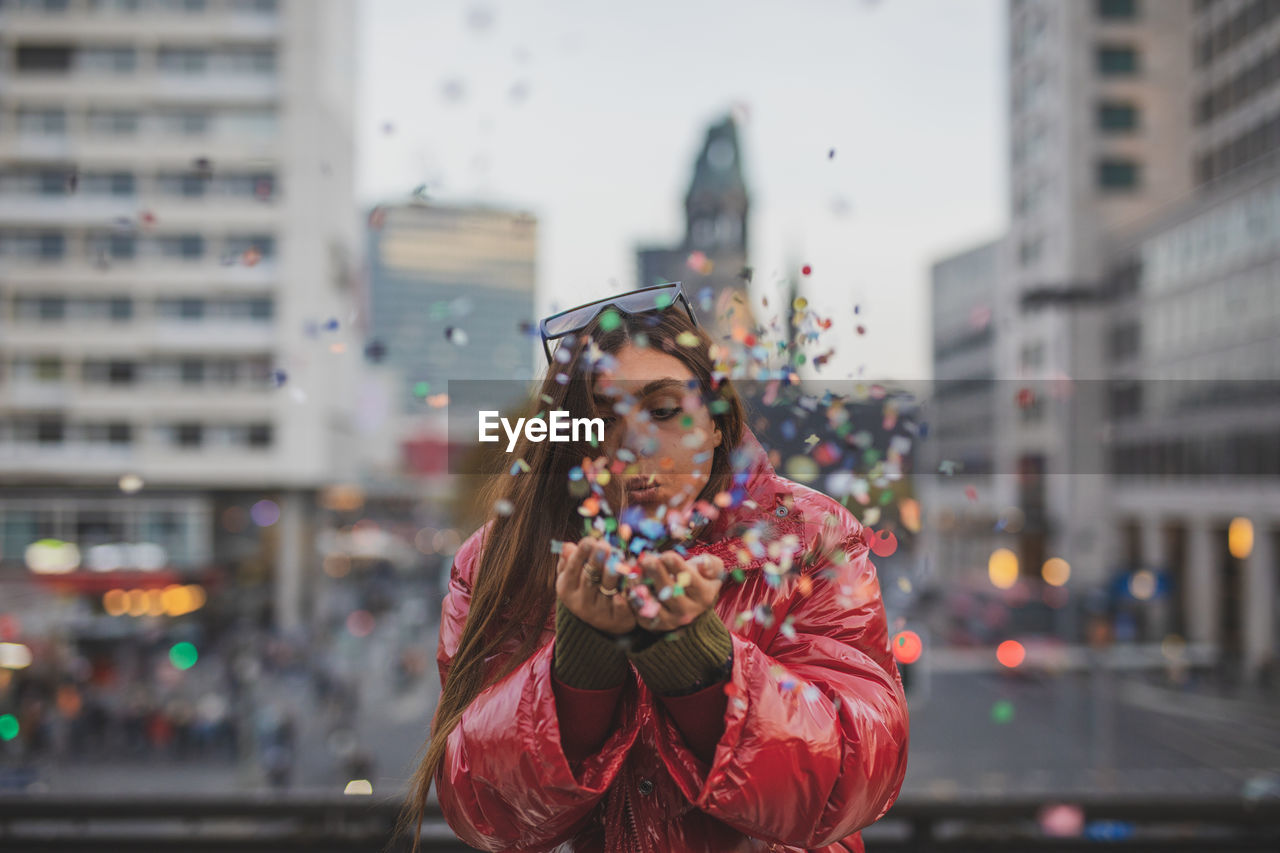 Young woman blowing colorful confetti in city during sunset