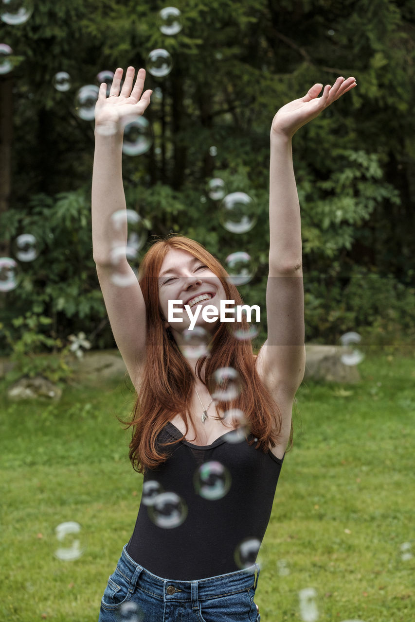 Happy woman with arms raised playing amidst soap bubbles in yard