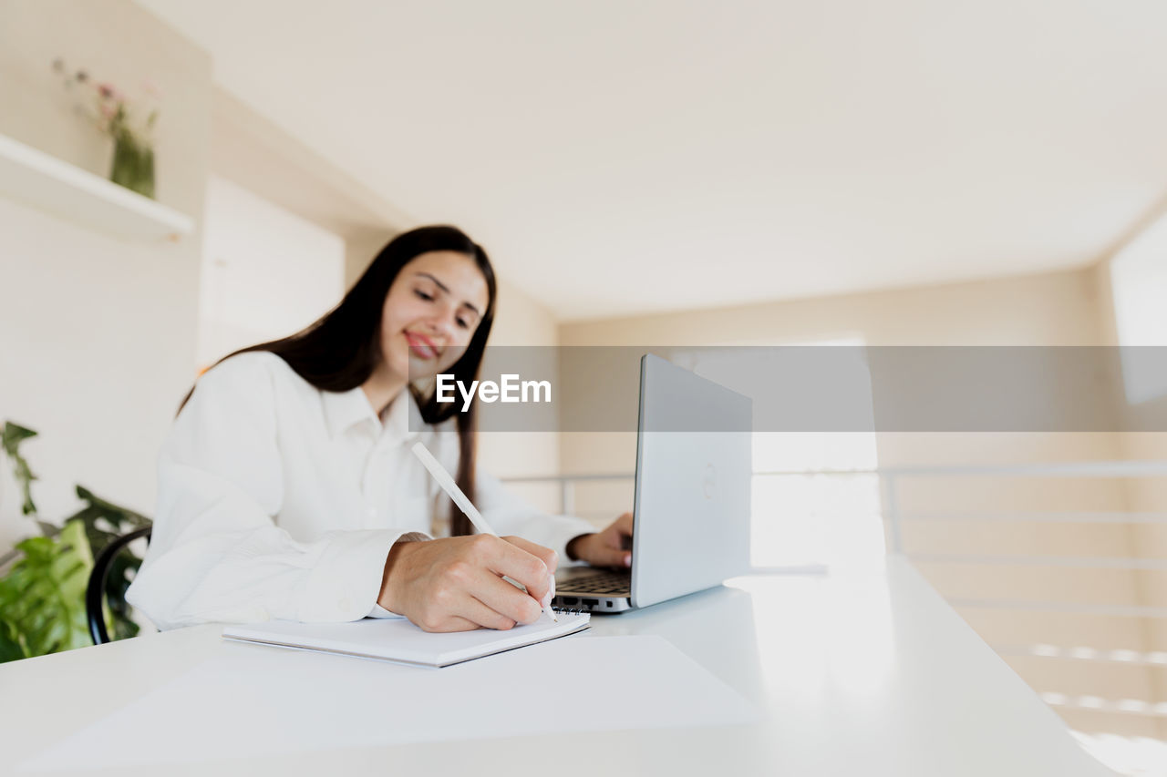 Caucasian young business woman sitting at desk, reading from computer, taking notes at notebook