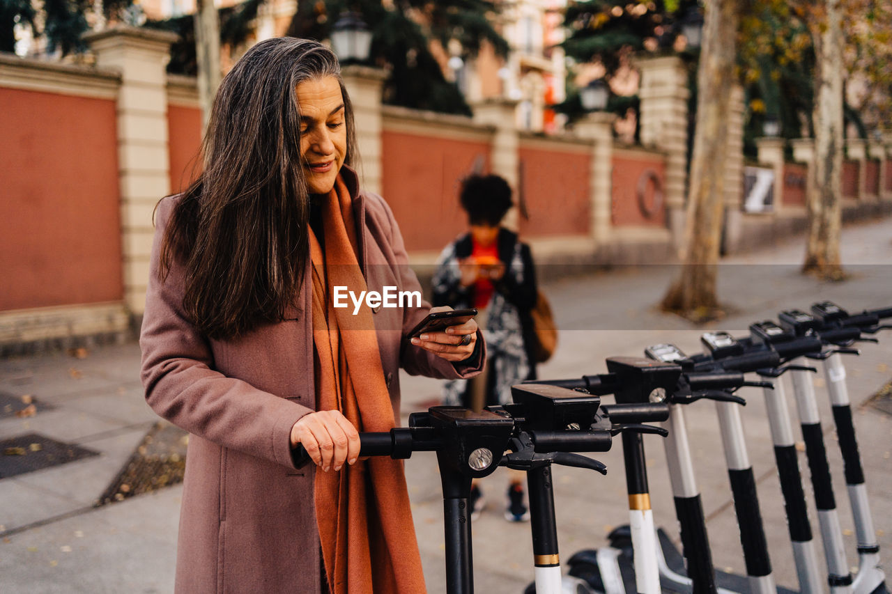 Hispanic long haired lady in warm coat standing on city street and doing contactless payment for electric scooter with smartphone using application