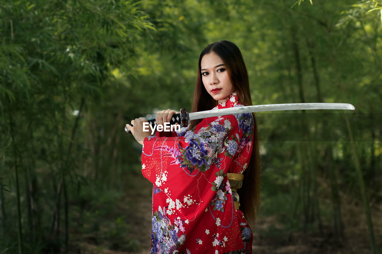 Portrait of young woman holding sword while standing in forest