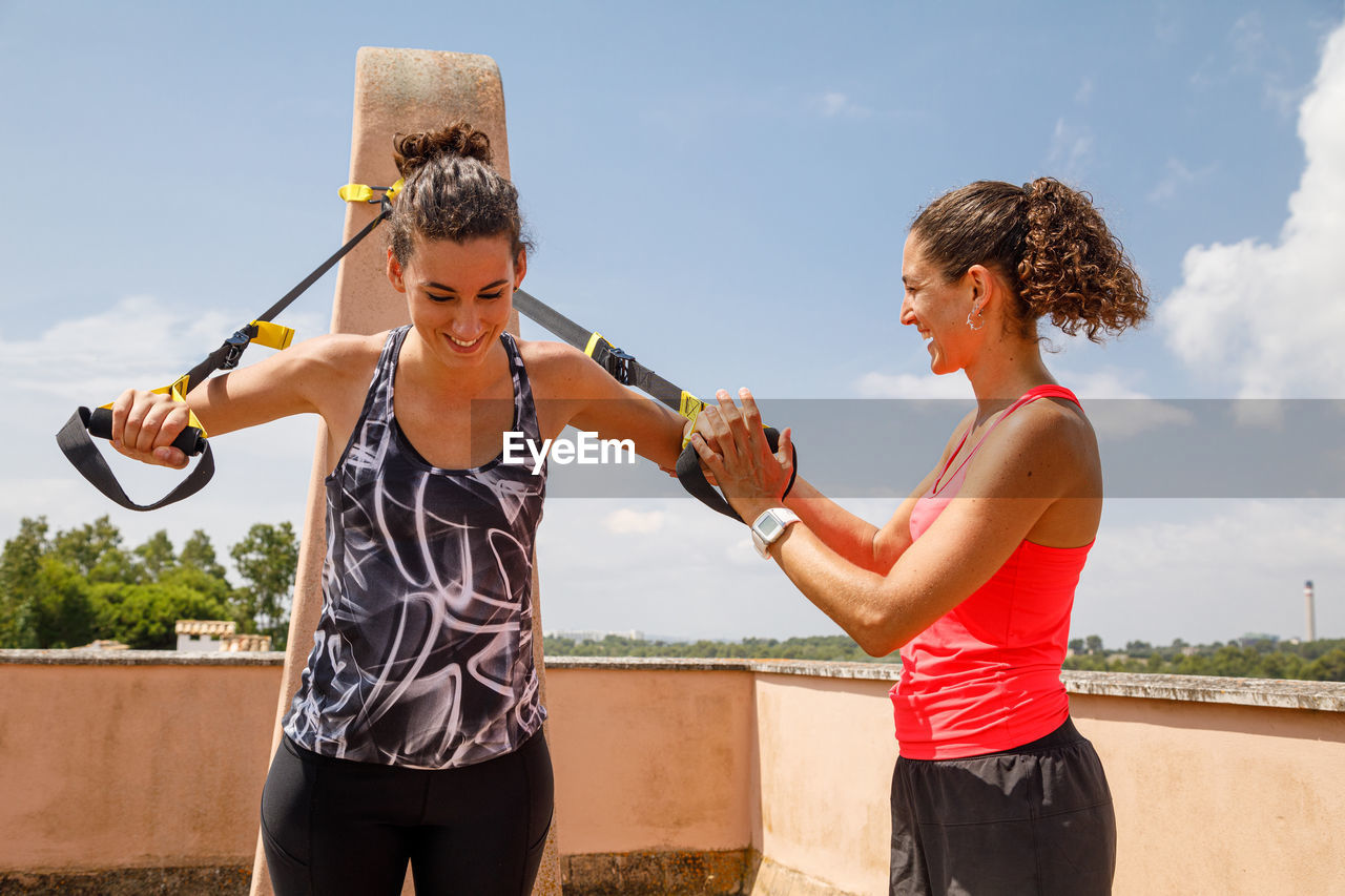 Expressive female trainer supporting concentrated sportswoman doing exercises with trx straps during training on terrace in summer