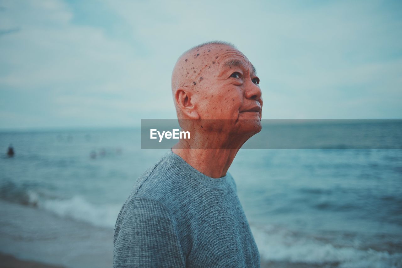 Thoughtful senior man standing at beach against sky during sunset