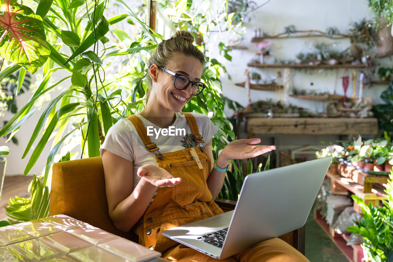 Smiling woman video conferencing over laptop while sitting amidst plants at home