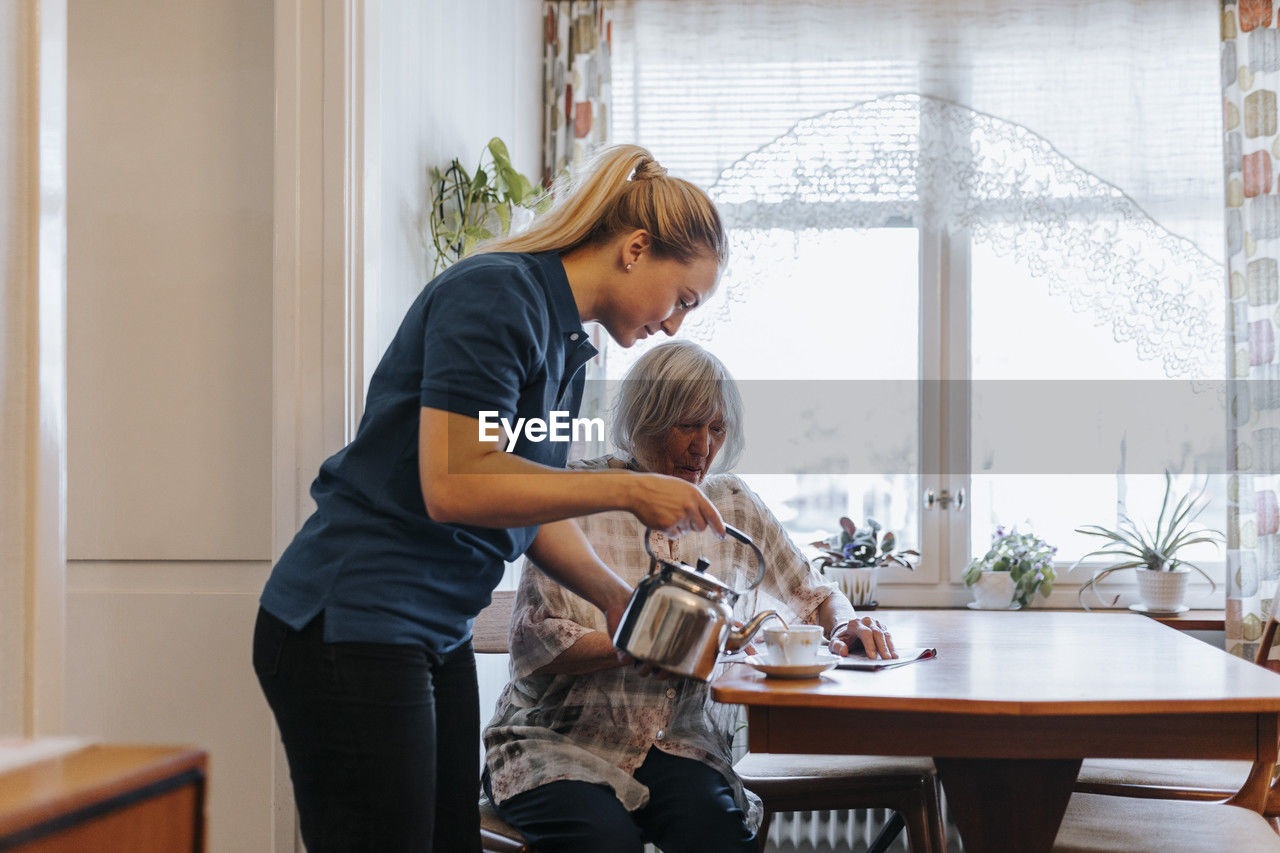 Side view of female caregiver serving tea to senior woman sitting at dining table