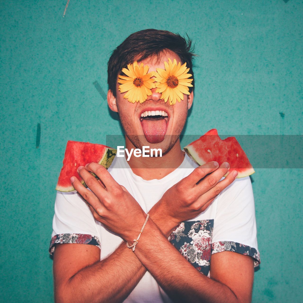 Man holding watermelon slices with eyes covered by flowers against wall