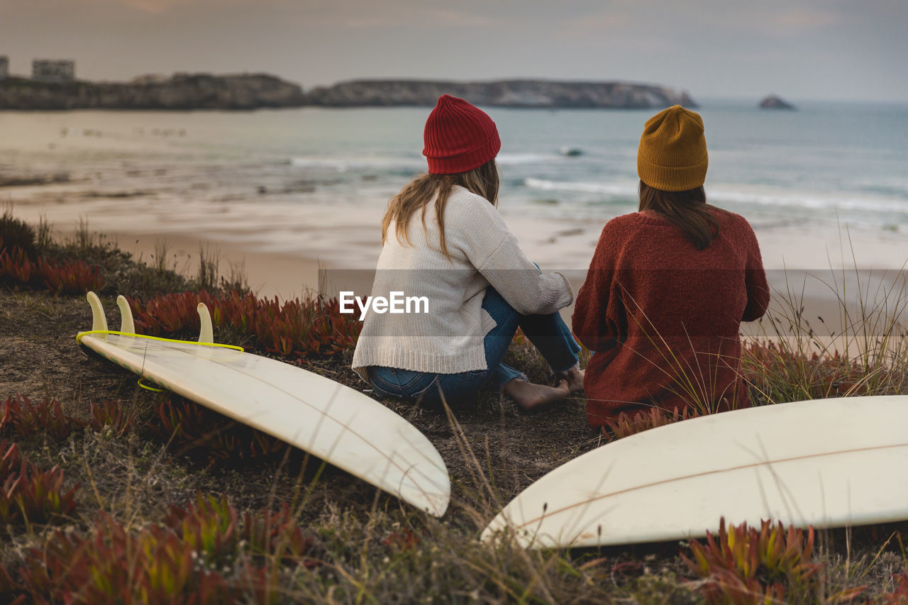 Rear view of women sitting by surfboard at beach