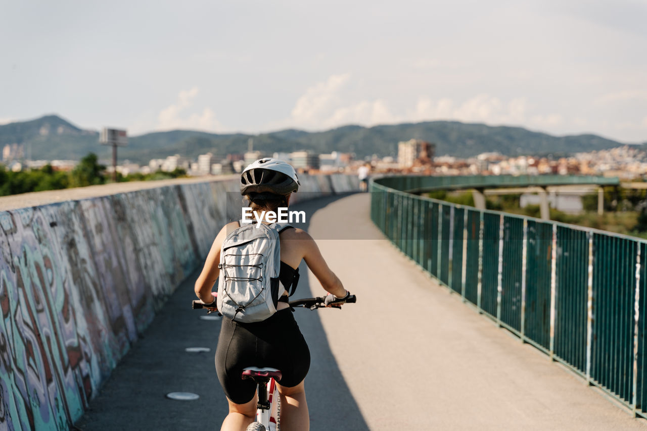 Back view confident young female bicyclist in sportswear and helmet riding bike on fenced curvy paved track while training alone in summer day