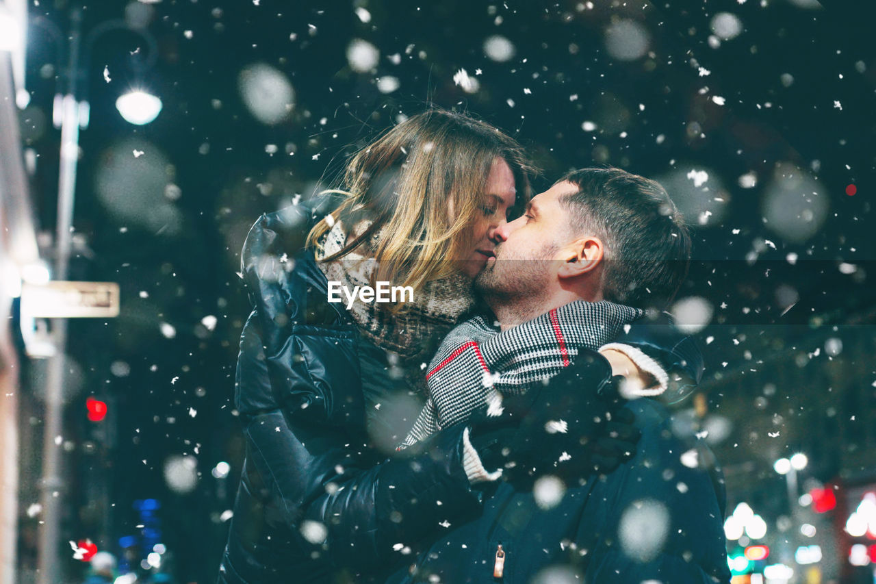 Couple romancing while standing against sky at night during snowing