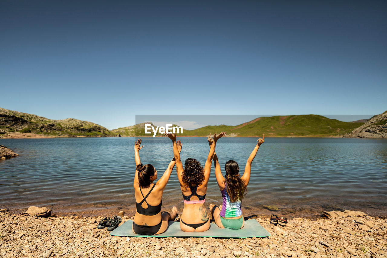 Back view of group of female friends in swimwear having fun and raising arms while enjoying summer journey on shore of mountain lake in pyrenees