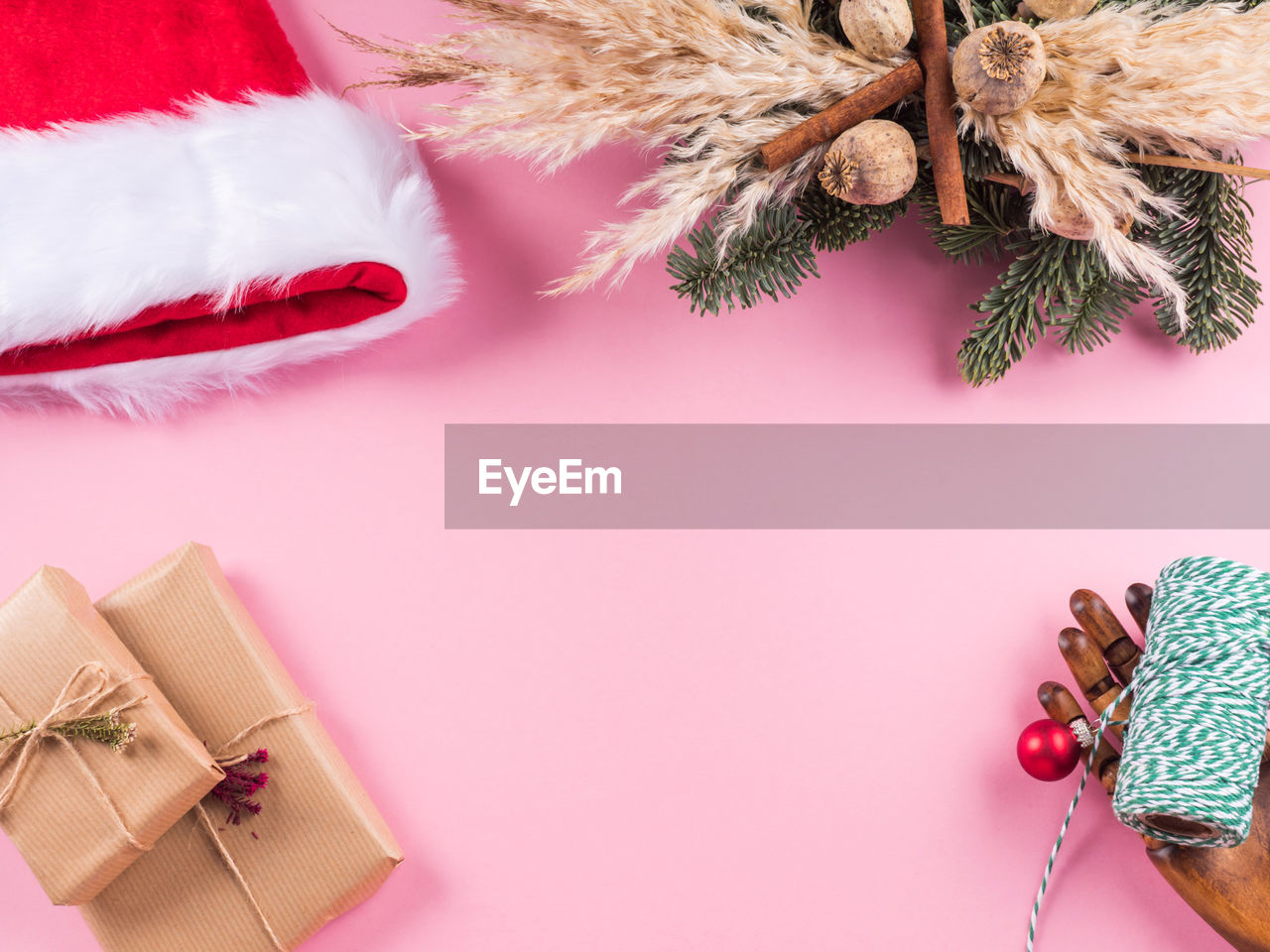 Red santa claus cap, spruce and pampas wreath, wrapped christmas presents on pink background. 