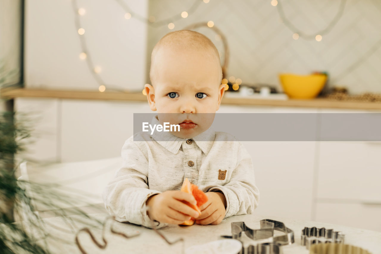 A sad kid with a grapefruit in his hands is sitting at a table with cookie cutters on it.