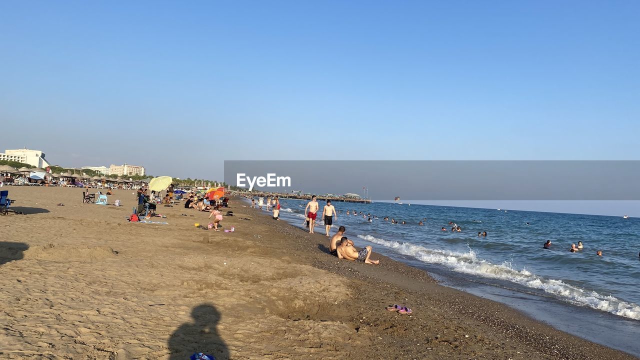 beach, sea, land, body of water, water, sky, coast, sand, large group of people, group of people, nature, ocean, crowd, shore, holiday, vacation, trip, clear sky, horizon over water, horizon, leisure activity, sunny, day, men, lifestyles, travel destinations, travel, scenics - nature, beauty in nature, summer, sunlight, women, adult, tourism, wave, blue, copy space, outdoors, coastline, tourist, relaxation, motion, sports, weekend activities