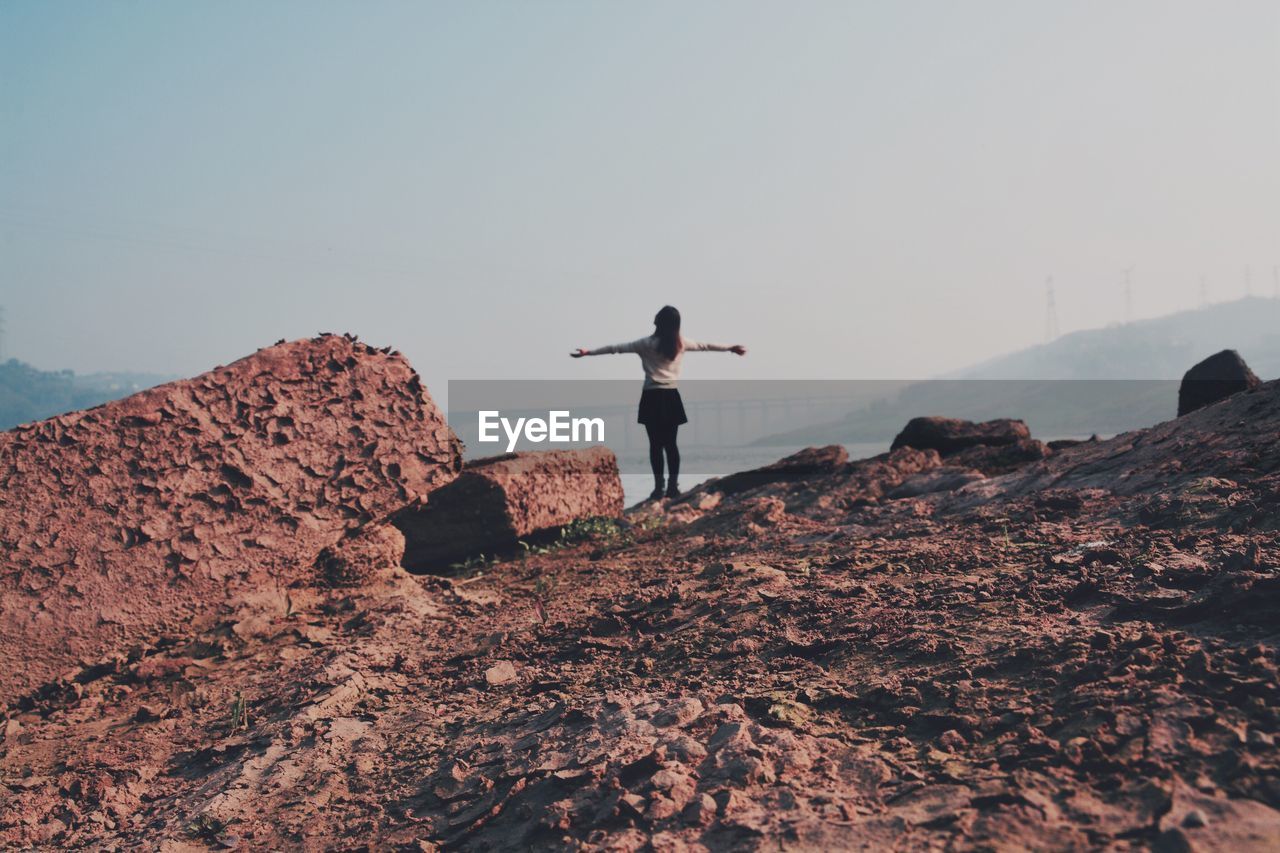 Rear view of woman with arms outstretched standing on rock formation against sky