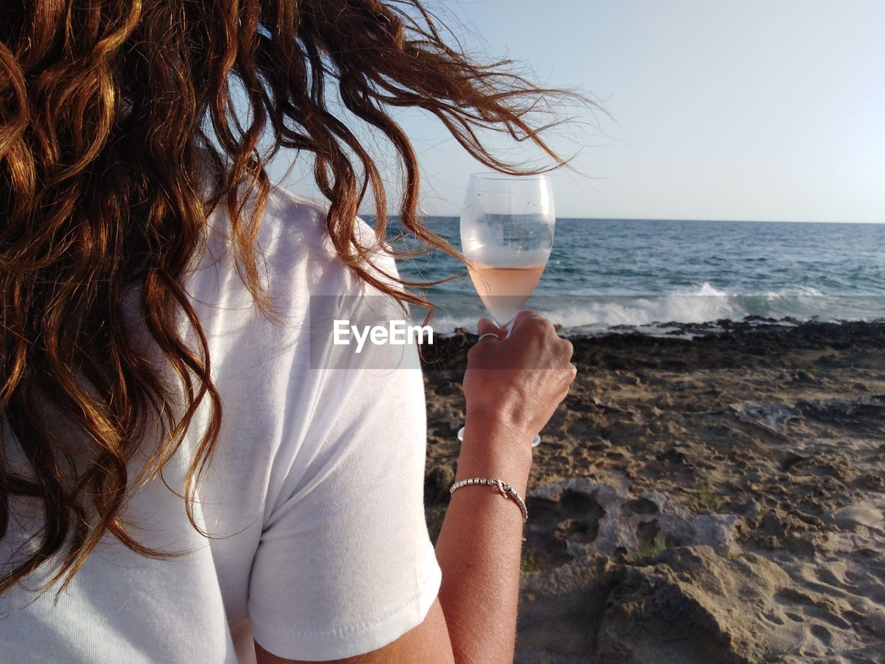 Midsection of woman holding wineglass while standing at beach against clear sky
