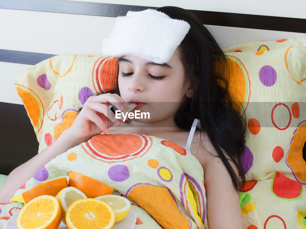 Teenage girl holding thermometer in mouth while lying on bed at home