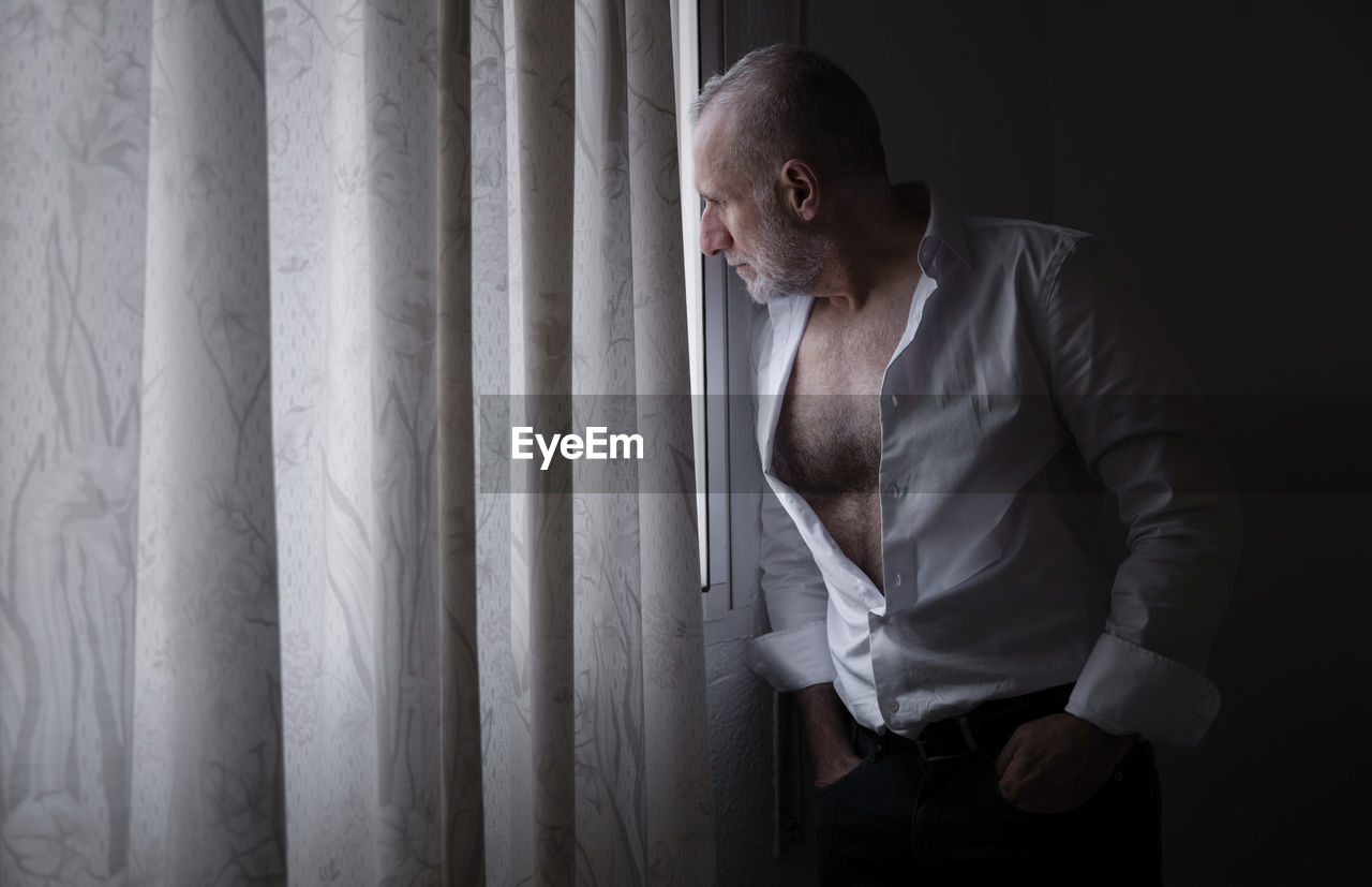 Adult man in white shirt in room looking out window