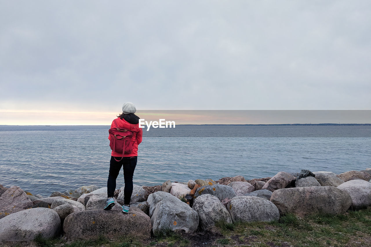 Woman in red jacket standing on rocks looking out over ocean horizon
