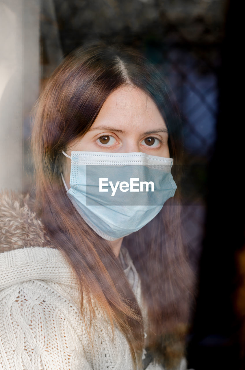 Young woman on self-isolation in a medical mask looks out the window