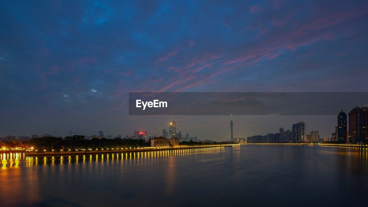 Illuminated buildings by river against sky at sunset