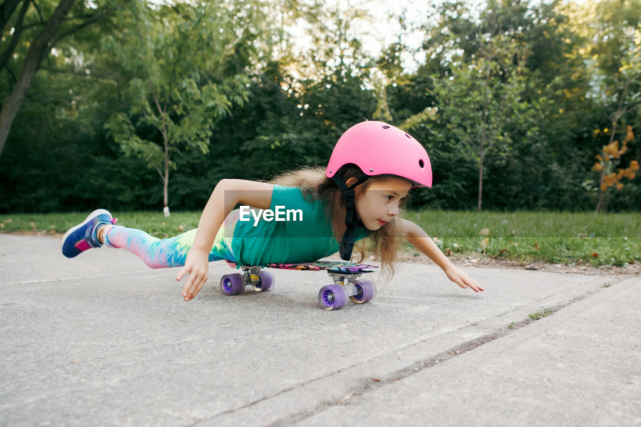 Happy smiling caucasian girl in pink helmet lying on tummy riding skateboard on road in park 